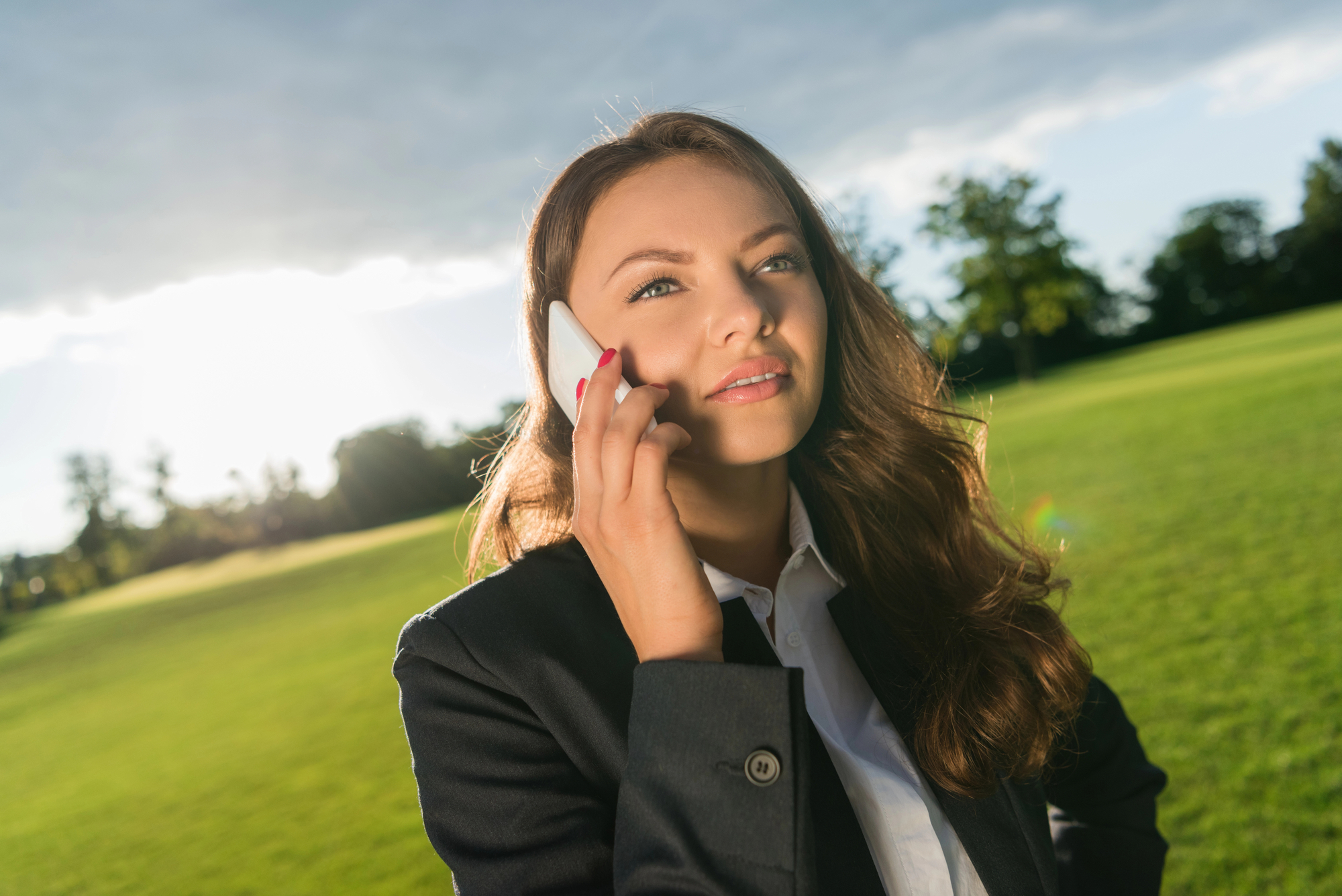 A woman in a dark blazer stands on a sunny lawn with trees in the background. She is holding a smartphone to her ear and looking thoughtfully into the distance. The sun is partially obscured by clouds, casting a soft light around her.