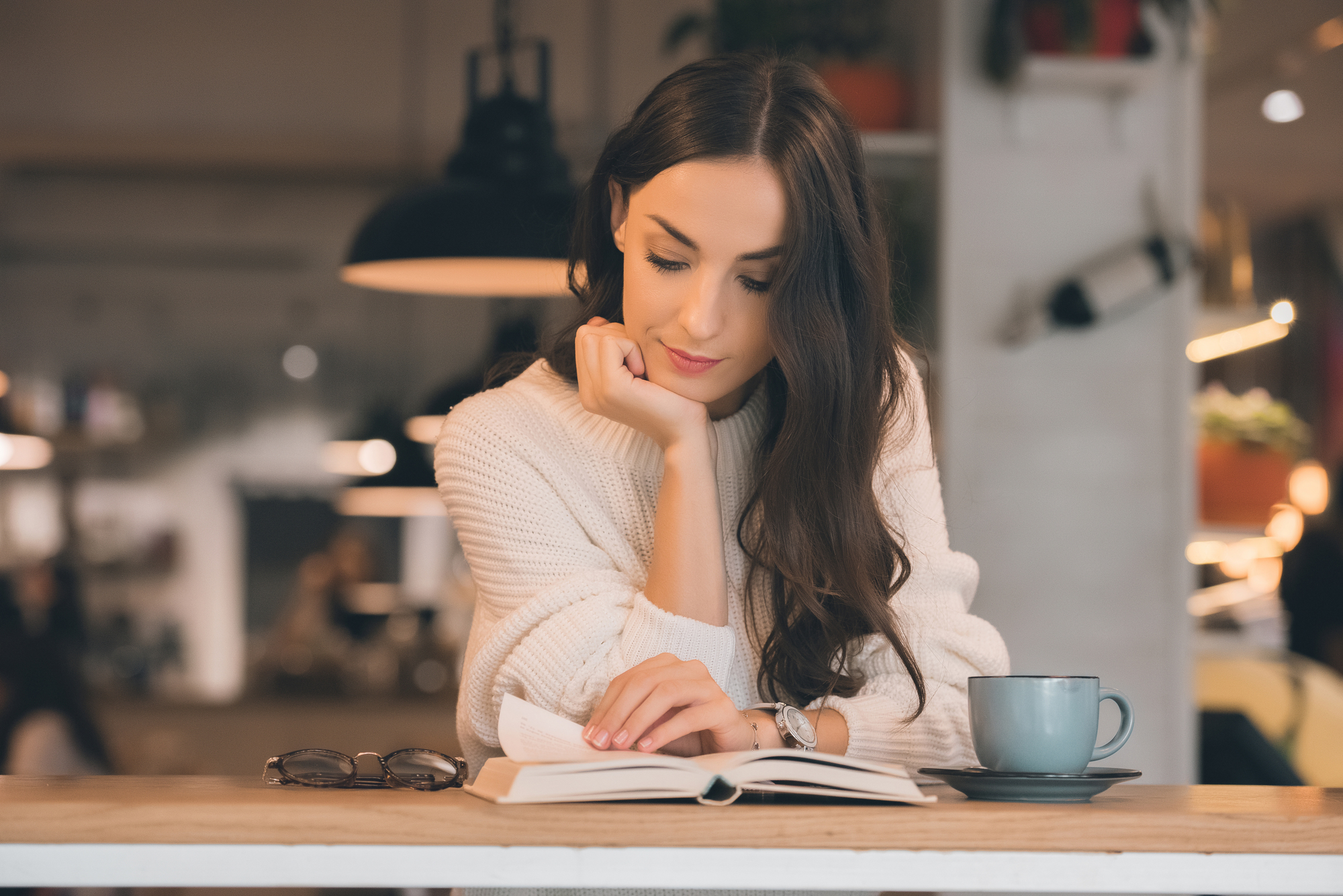 A woman with long dark hair sits at a wooden table in a cozy café, intently reading a book. She rests her chin on her hand, wearing a white sweater. A pair of glasses and a blue cup are on the table to her right. The background is softly lit and blurred.