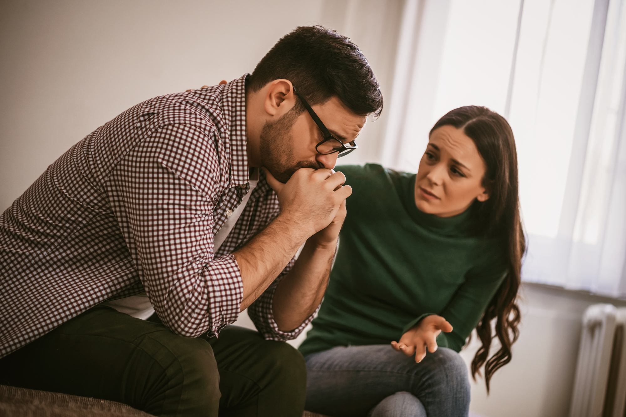 A man wearing glasses and a checkered shirt sits with his hands clasped under his chin, appearing distressed. A woman in a green sweater sits beside him, looking concerned and gesturing with her hand. They are indoors with a window in the background.