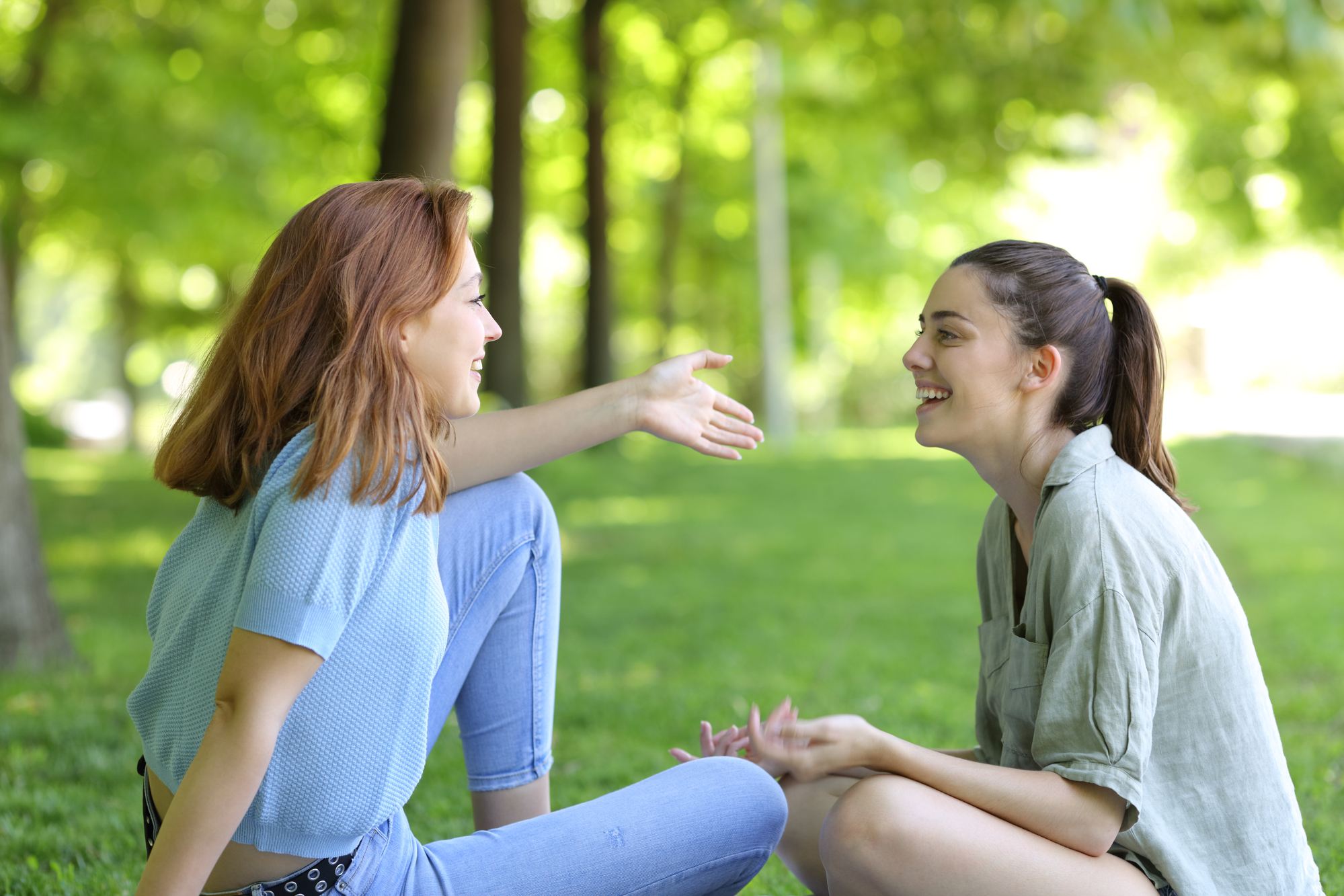 Two women are sitting on the grass in a park, engaged in a lively conversation. One woman with shoulder-length red hair is gesturing with her hand, while the other with dark hair in a ponytail is smiling, both appearing relaxed and happy. Trees are visible in the background.