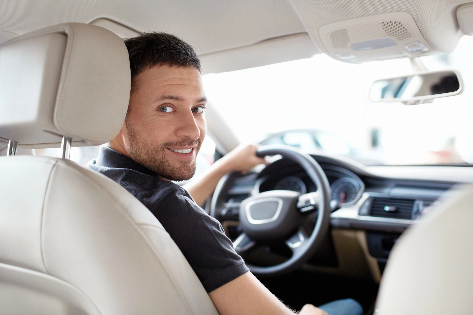 A man with short dark hair and a beard sits in the driver's seat of a car, smiling and looking back over his shoulder. The interior of the car is mostly light-colored, and the dashboard and steering wheel are visible.