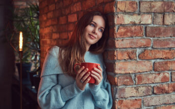 A woman with long brown hair, dressed in a light blue sweater, leans against a brick wall, smiling and gazing off into the distance. She holds a red mug with both hands and appears to be in a cozy, dimly lit room with a warm atmosphere.