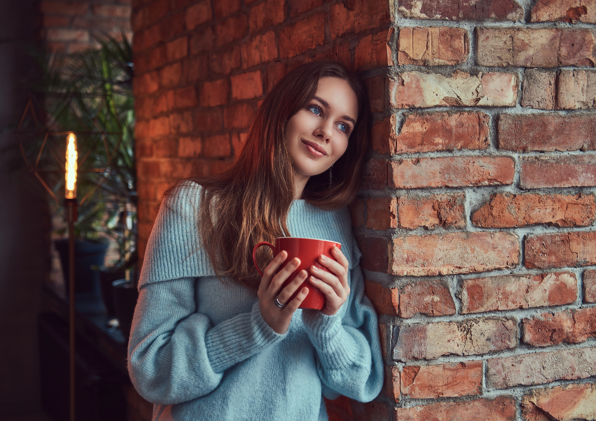 A woman with long brown hair, dressed in a light blue sweater, leans against a brick wall, smiling and gazing off into the distance. She holds a red mug with both hands and appears to be in a cozy, dimly lit room with a warm atmosphere.