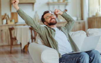 A bearded man wearing glasses and a green shirt sits on a couch with a laptop on his lap. He is stretching his arms overhead and smiling, appearing relaxed and happy. Behind him is a cozy, well-lit room with wooden furniture and decor.