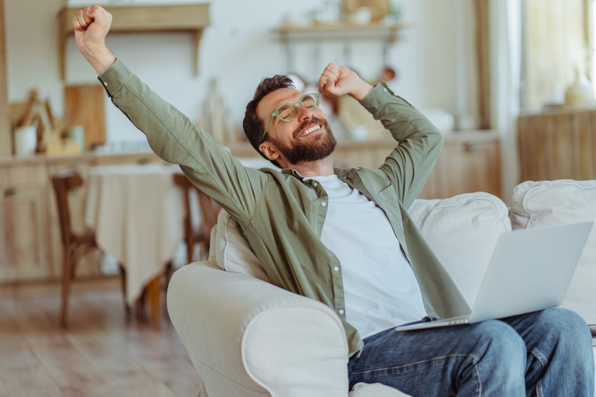 A bearded man wearing glasses and a green shirt sits on a couch with a laptop on his lap. He is stretching his arms overhead and smiling, appearing relaxed and happy. Behind him is a cozy, well-lit room with wooden furniture and decor.