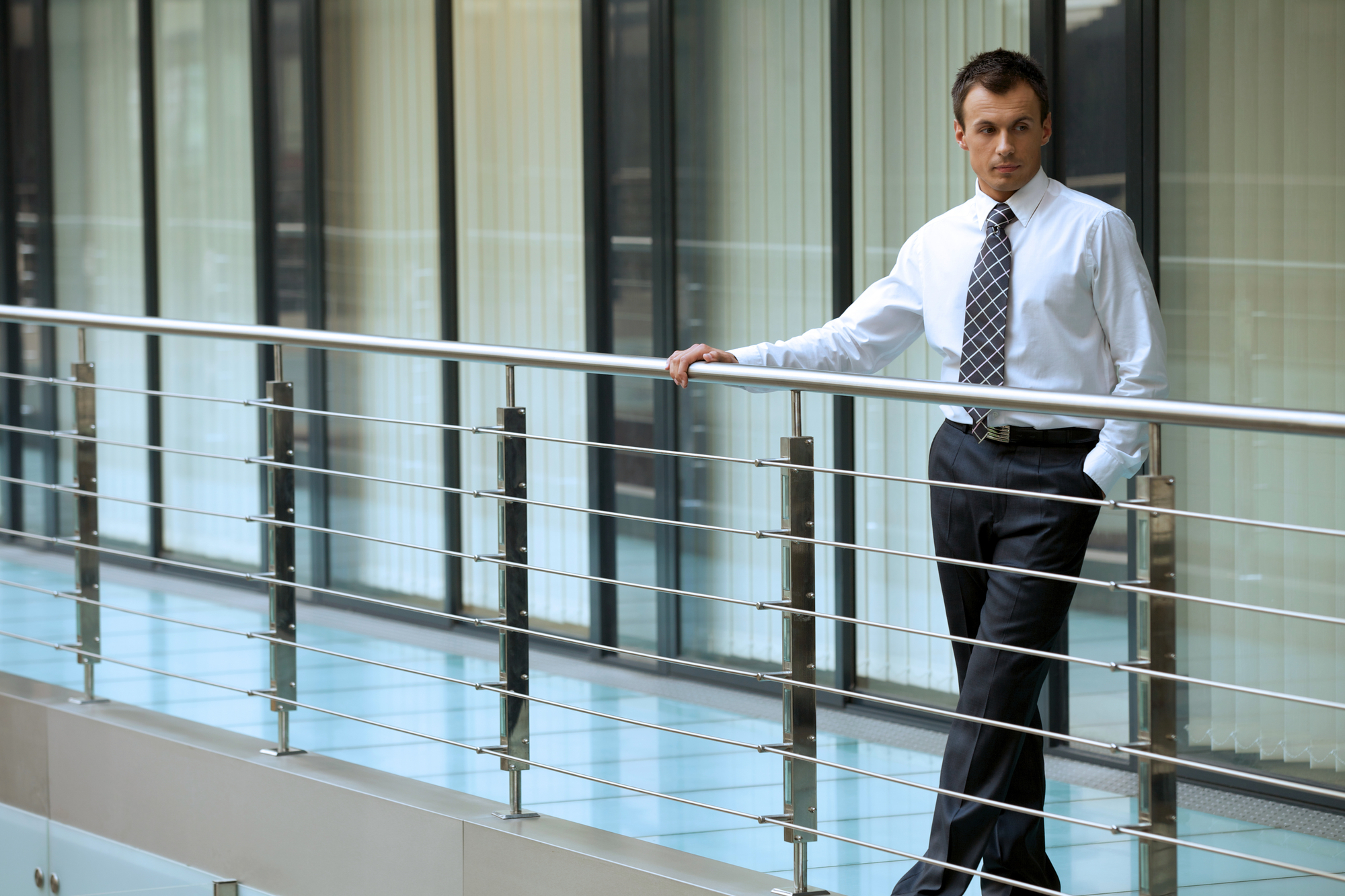 A man in business attire stands on a modern walkway with metal railings, gazing to his right. He is wearing a light blue dress shirt, dark trousers, and a patterned tie. The background features large glass windows with vertical blinds, reflecting a professional environment.