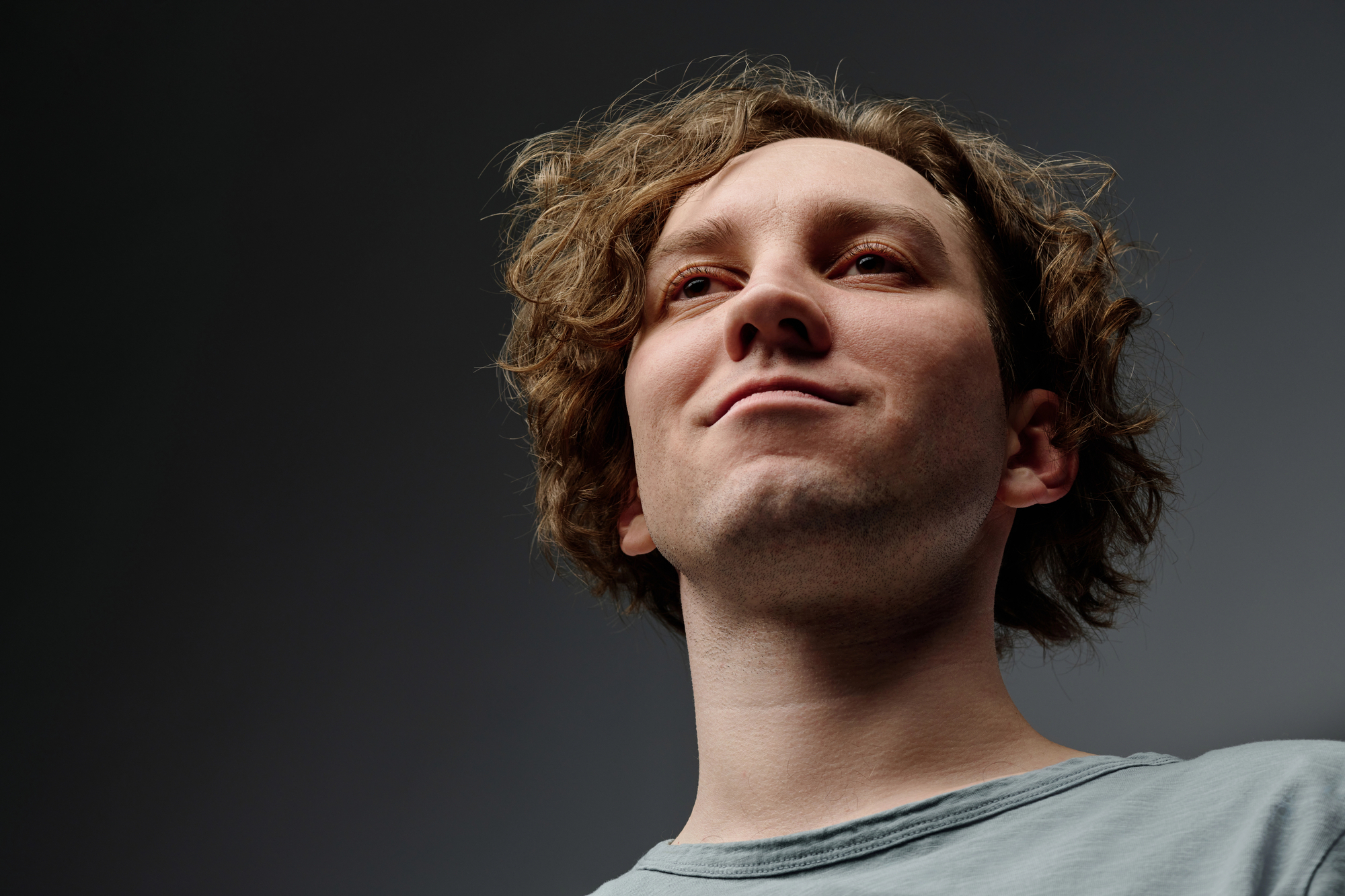 A close-up, low-angle shot of a young man with wavy, medium-length hair looking slightly upward. He is wearing a light-colored shirt and the background is a gradient of dark and lighter shades of grey, giving a dramatic effect.