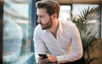 A man with a beard and short hair, wearing a white dress shirt, looks thoughtfully out of a window while holding a smartphone. He is leaning on a window ledge in an indoor setting with a plant in the background.