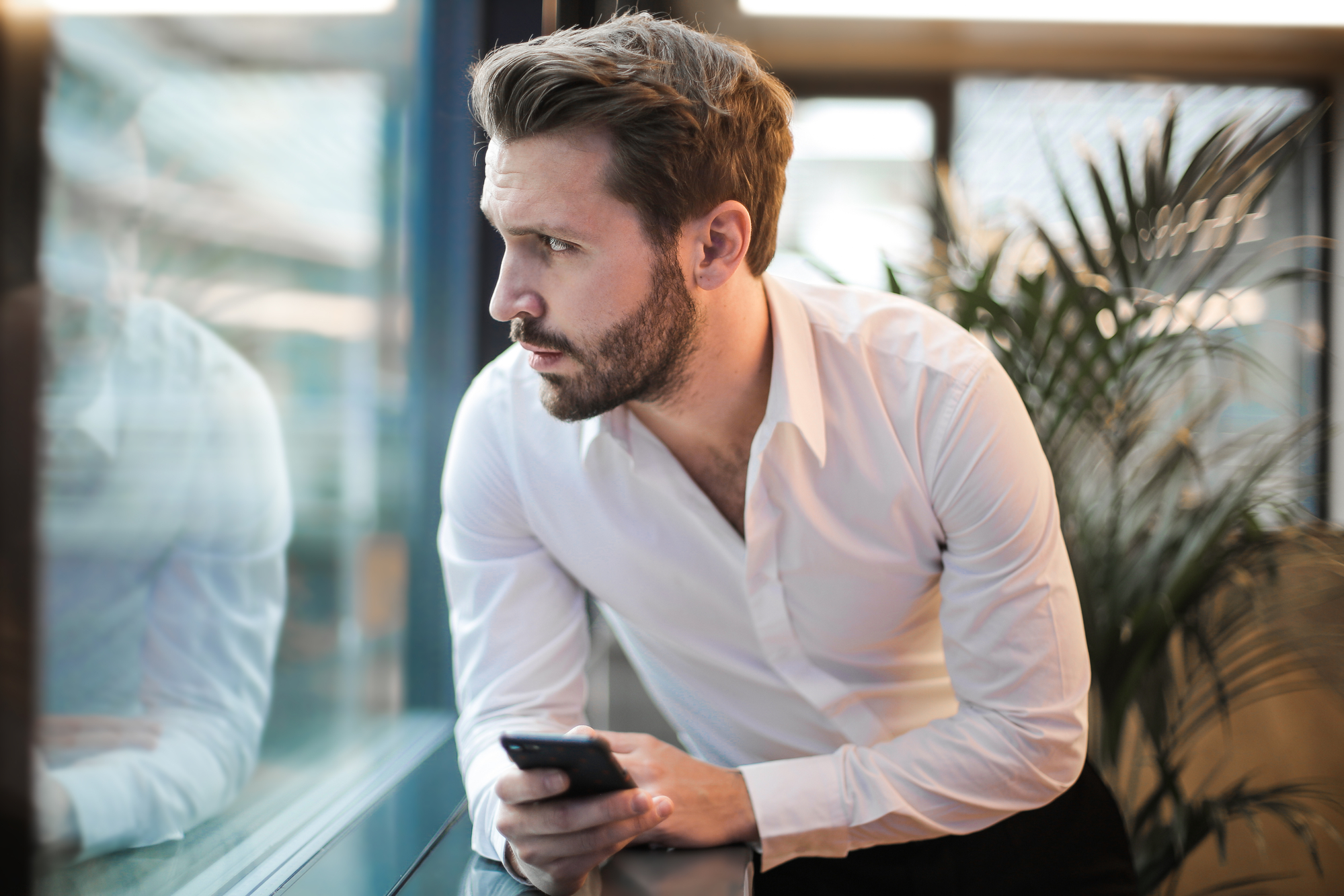 A man with a beard and short hair, wearing a white dress shirt, looks thoughtfully out of a window while holding a smartphone. He is leaning on a window ledge in an indoor setting with a plant in the background.