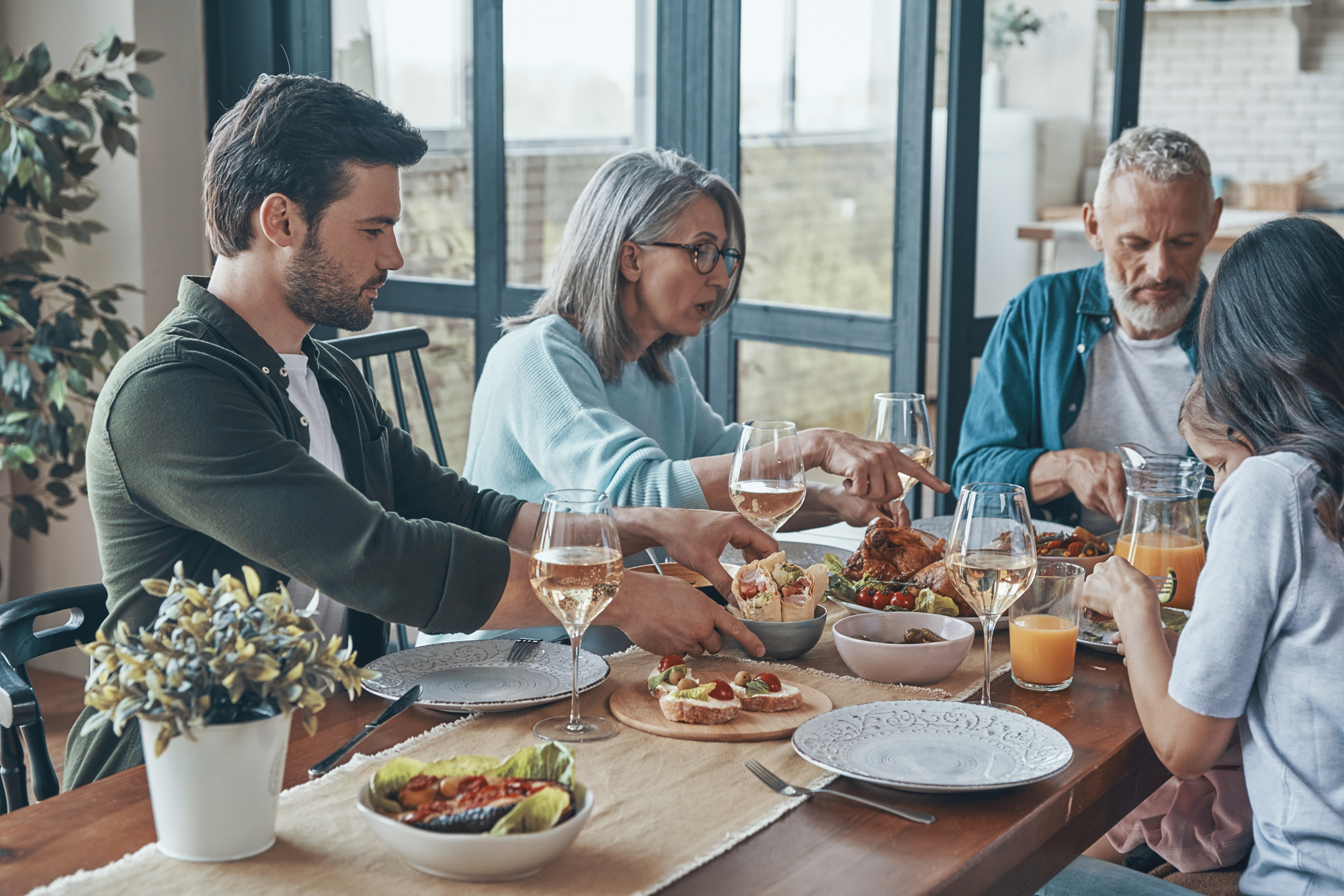 Four people are seated around a table, enjoying a meal together. The table is set with various dishes, including a salad, bread, and other foods. The setting appears to be a home with large windows in the background, allowing natural light to illuminate the scene.