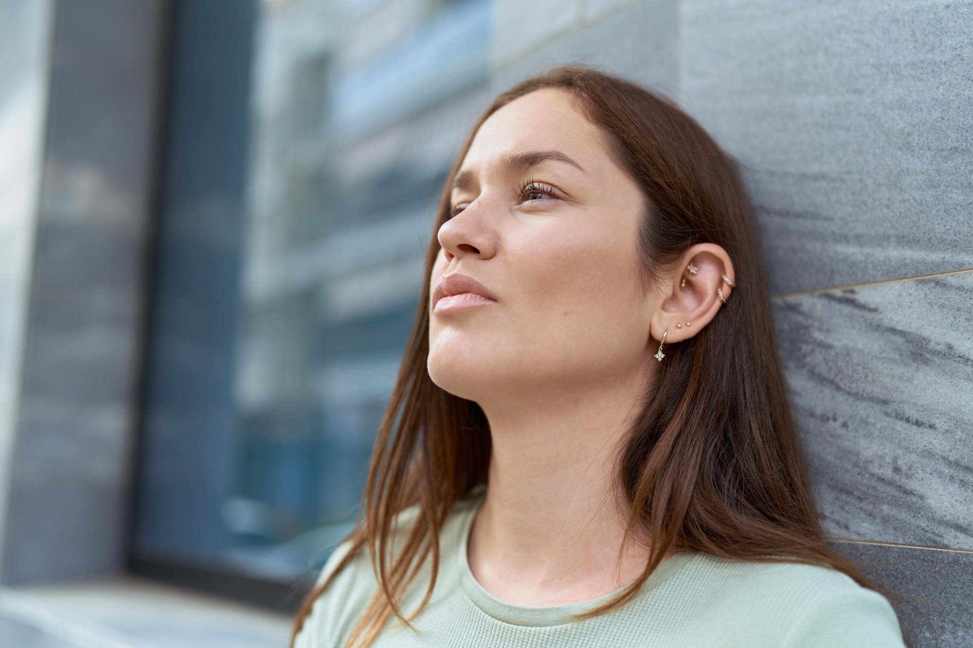 A woman with long brown hair, wearing a light green shirt, leans against a gray marble wall, looking slightly upward with a contemplative expression. She has multiple ear piercings and is outdoors with a blurred urban background.