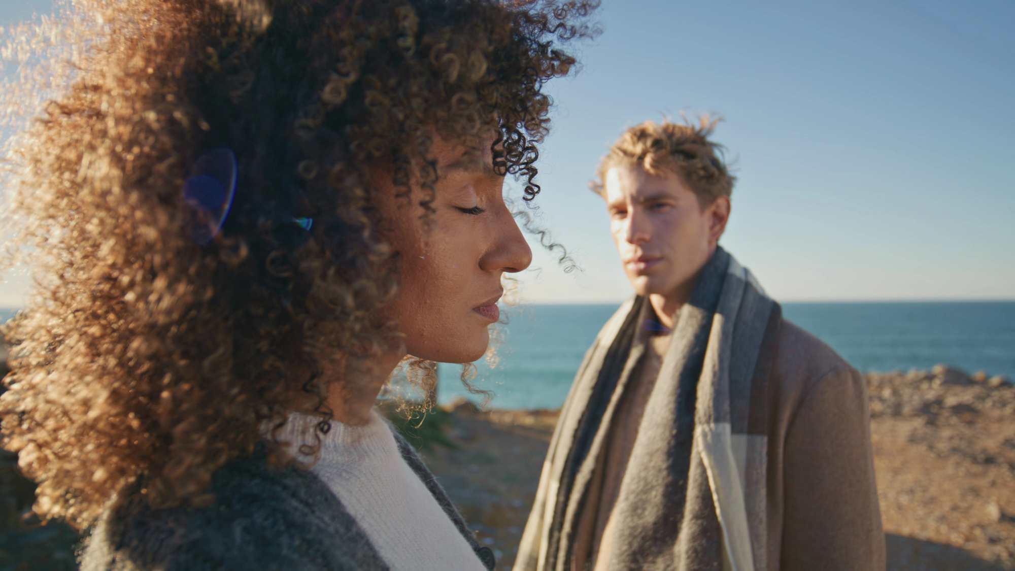 Two people stand near the ocean on a sunny day. The person in the foreground with curly hair, eyes closed, wears a gray sweater. The person in the background, with light hair and a scarf, looks at the first person. The sea and clear sky are visible behind them.