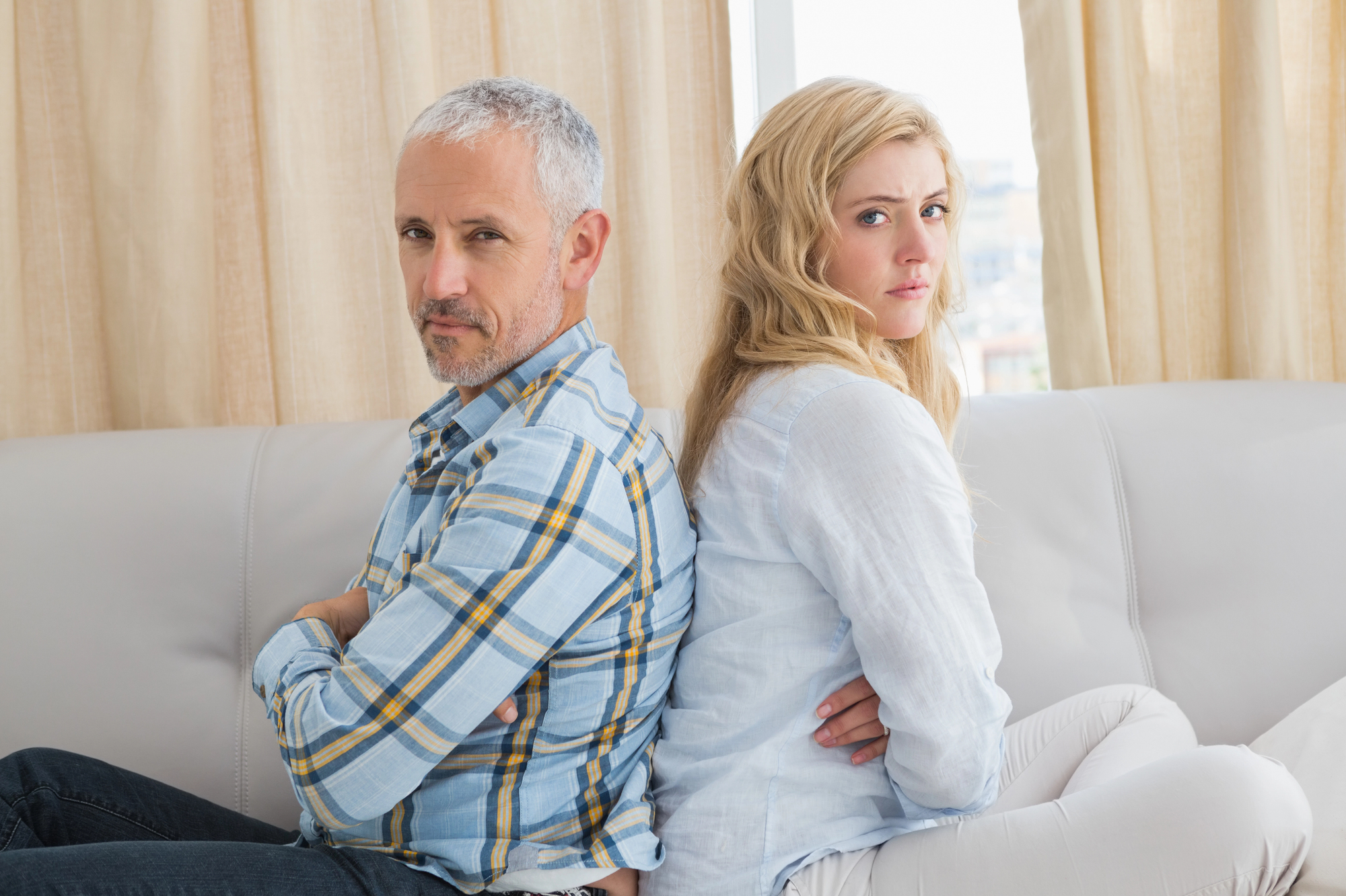 A man with short gray hair and a plaid shirt sits back-to-back with a woman with long blonde hair and a white top on a sofa. Both have their arms crossed and appear to be unhappy or upset, facing away from each other. Beige curtains are in the background.