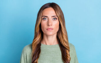 A woman with long, wavy brown hair featuring blonde highlights is wearing a light green sweater and standing against a light blue background. She has a serious expression and is looking directly at the camera.