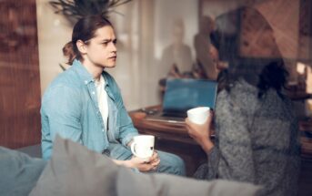 Two people are sitting on a couch in a cozy room, engaged in conversation while holding mugs. One person has light brown hair tied back and is wearing a blue shirt. A laptop is open on the table behind them. The setting appears casual and relaxed.