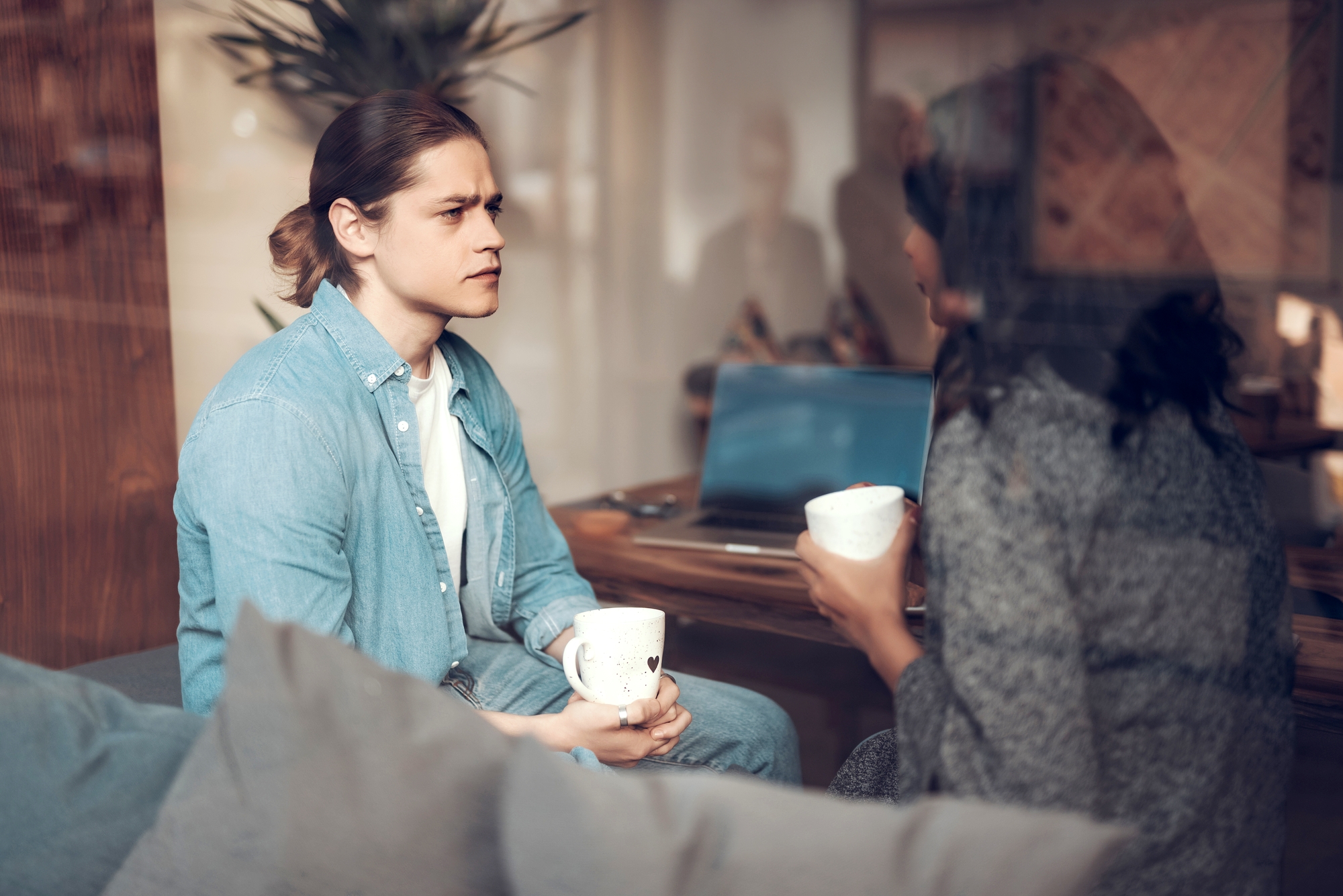 Two people are sitting on a couch in a cozy room, engaged in conversation while holding mugs. One person has light brown hair tied back and is wearing a blue shirt. A laptop is open on the table behind them. The setting appears casual and relaxed.