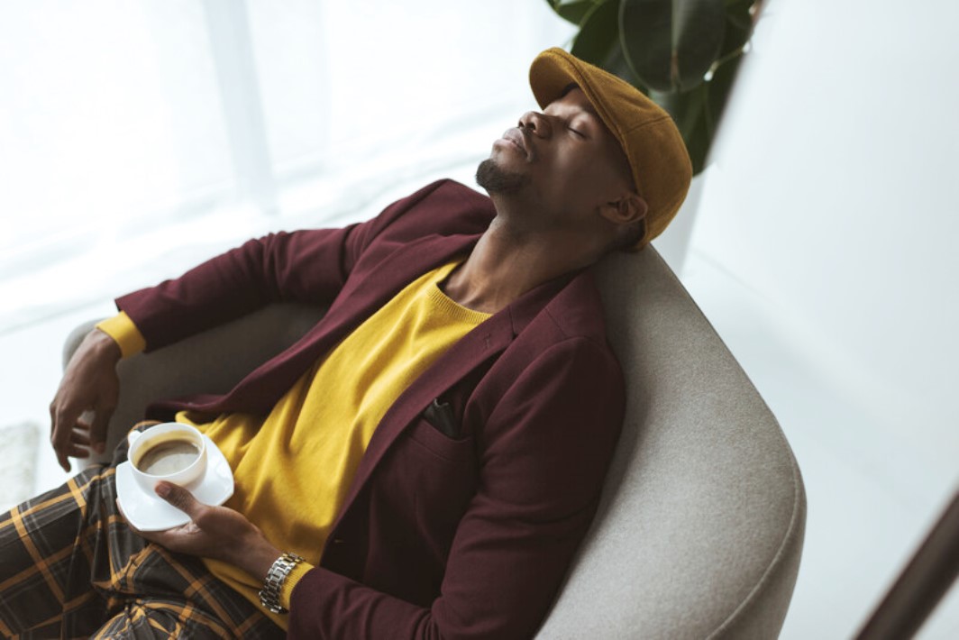 A man wearing a mustard yellow cap, bright yellow shirt, maroon blazer, and plaid pants is sitting in a gray chair with his eyes closed. He is holding a white coffee cup in his right hand and appears relaxed. Light filters through a window in the background.