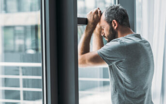 A man with short gray hair and a beard leans against a window, resting his forearms on the top of the frame. He is wearing a gray t-shirt and appears to be deep in thought or stressed. The background shows a blurred view of a modern exterior.