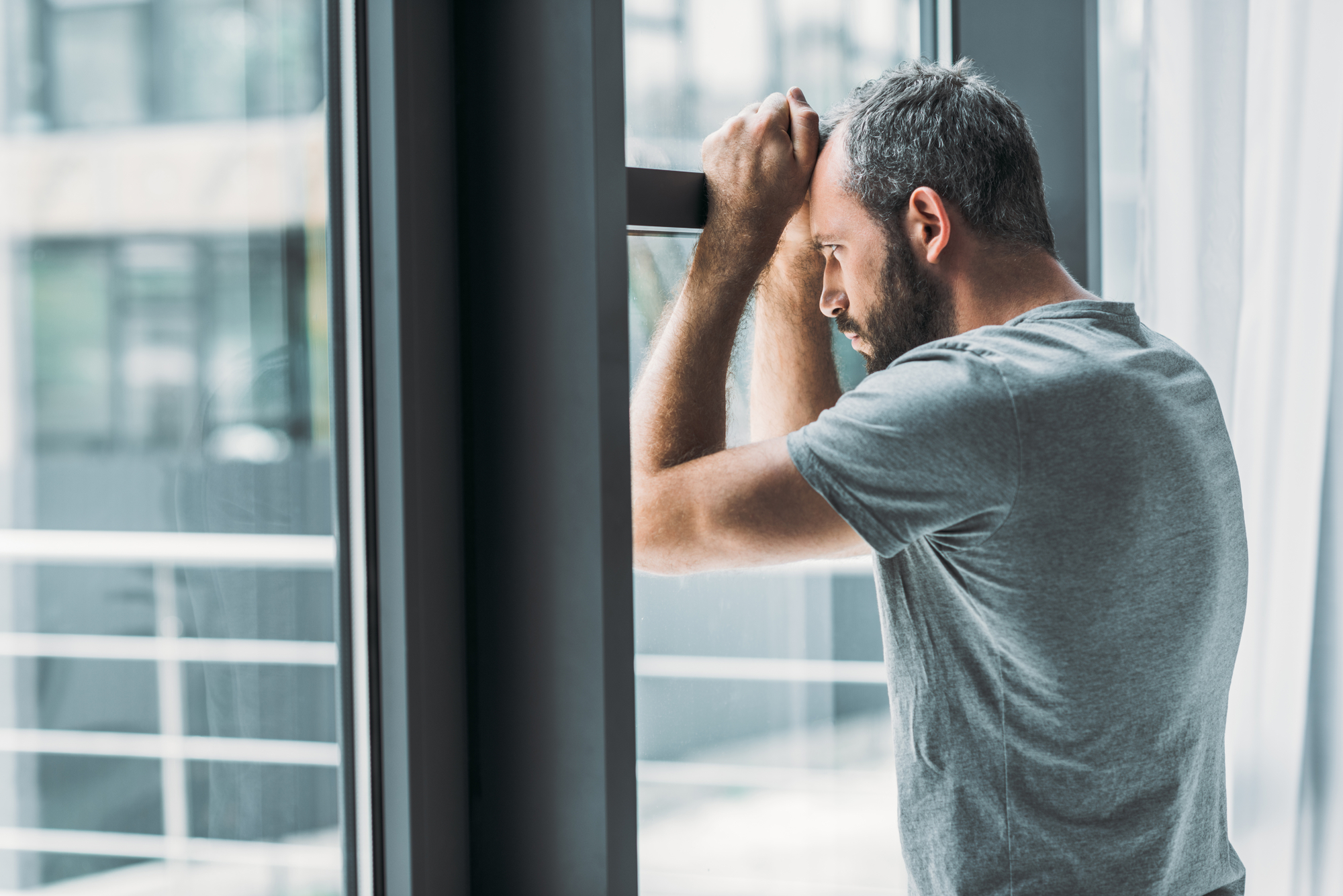 A man with short gray hair and a beard leans against a window, resting his forearms on the top of the frame. He is wearing a gray t-shirt and appears to be deep in thought or stressed. The background shows a blurred view of a modern exterior.