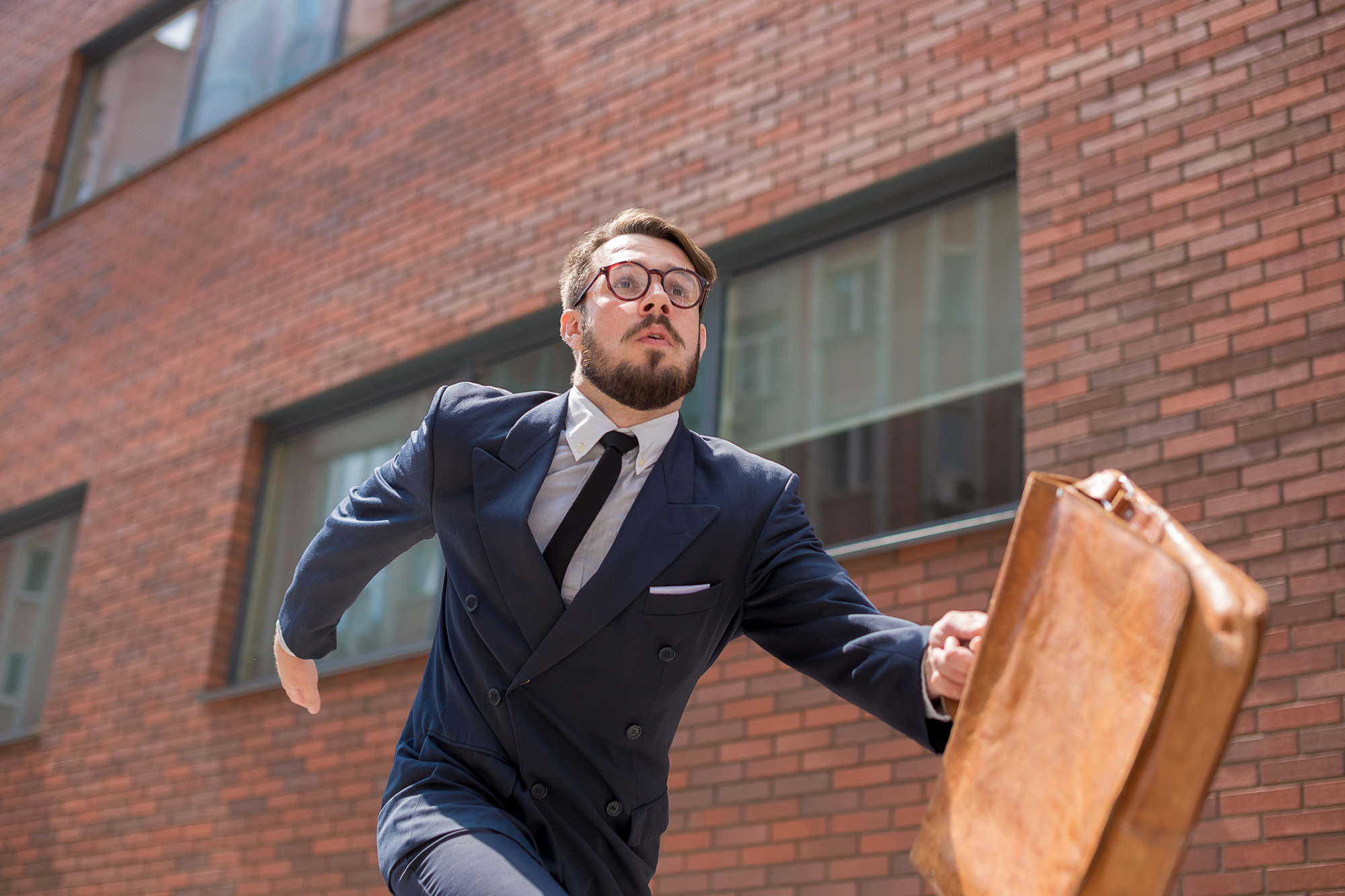 A man in a suit with a briefcase is running along a brick building. He has glasses, facial hair, and a determined expression. The scene suggests he might be in a hurry or late for something important.