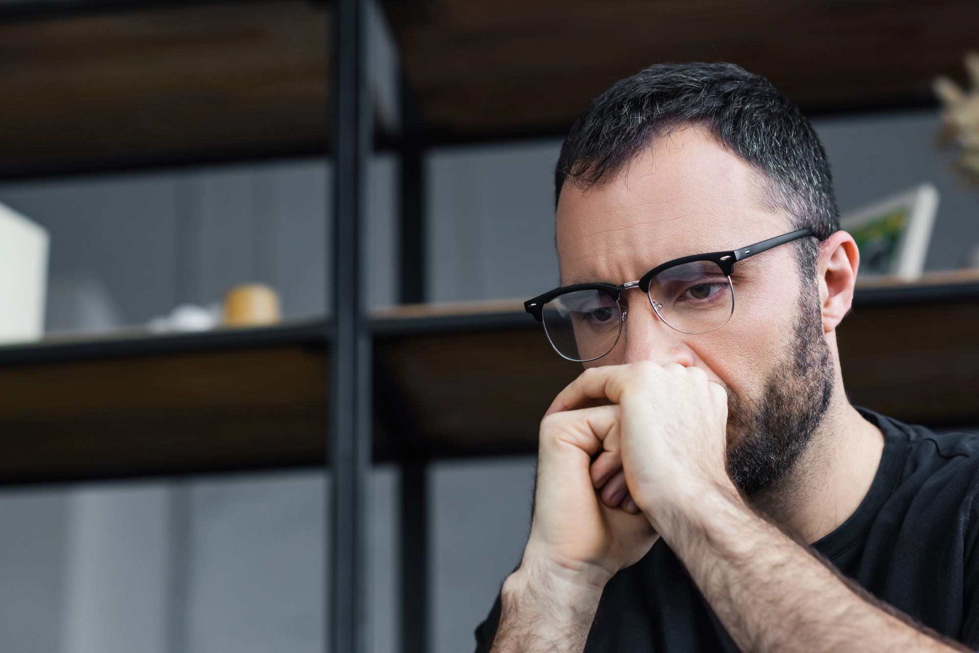 A man with short dark hair and glasses, wearing a black shirt, is sitting indoors, resting his chin on his hands with a pensive expression. There are shelves with various items in the blurry background.