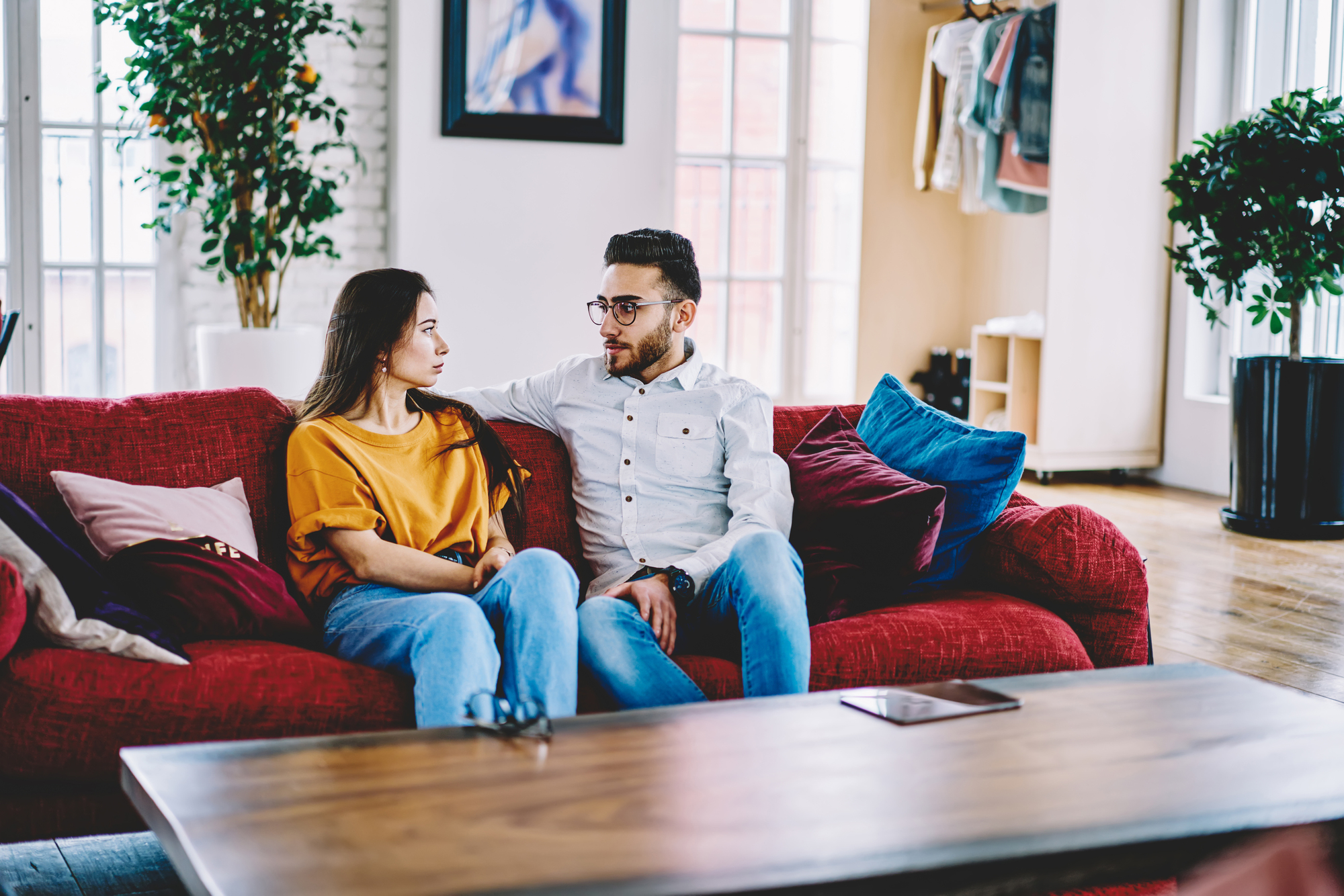 A man and a woman are sitting on a red sofa in a well-lit living room. The man, wearing glasses and a white shirt, has his arm around the woman, who is wearing a yellow top. They appear to be engaged in a serious conversation. A coffee table and plants are visible.