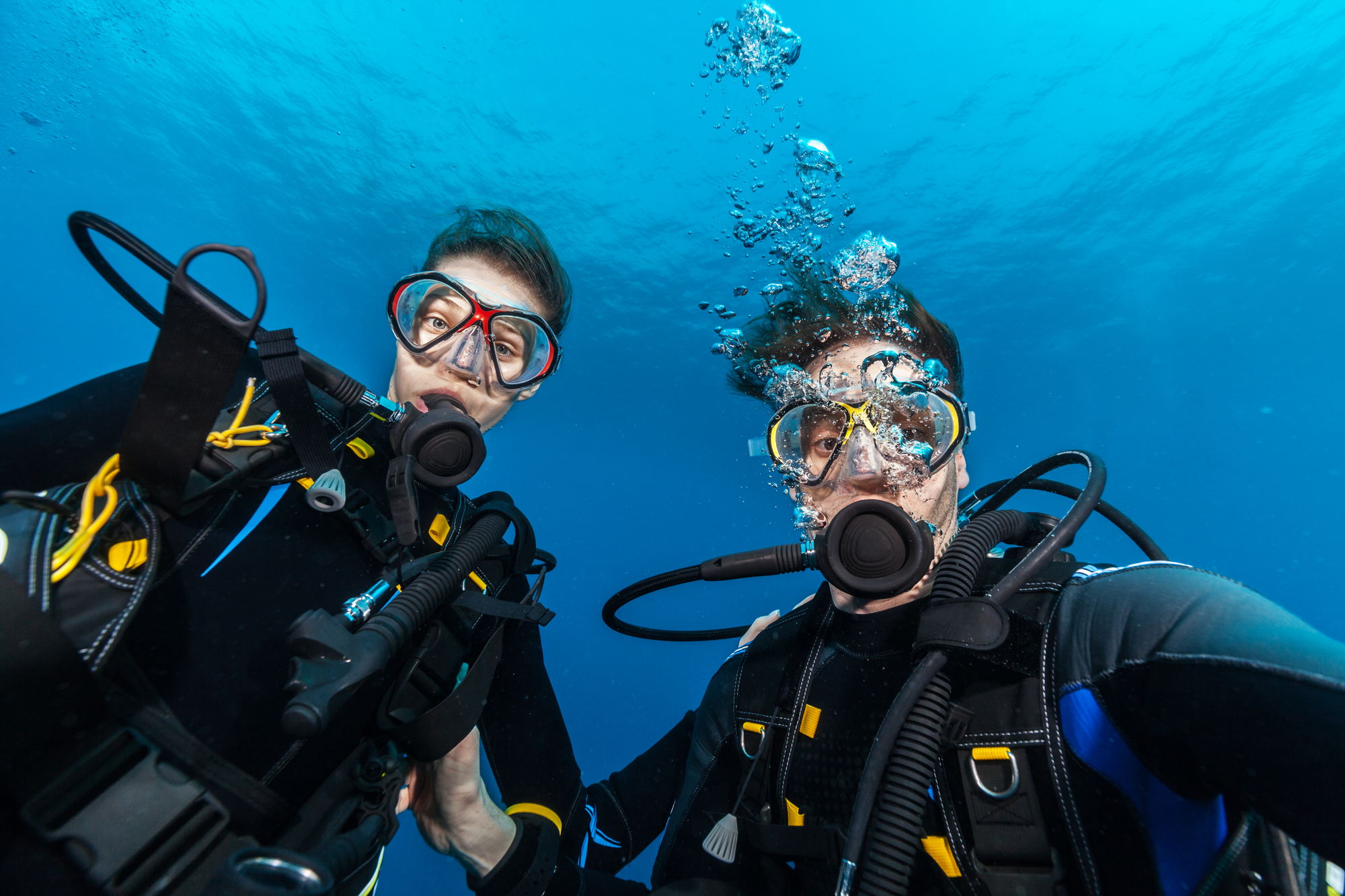 Two scuba divers underwater, wearing black wetsuits, masks, and mouthpieces, hold onto their equipment while looking into the camera. Bubbles rise to the surface, and the background features a deep blue ocean.