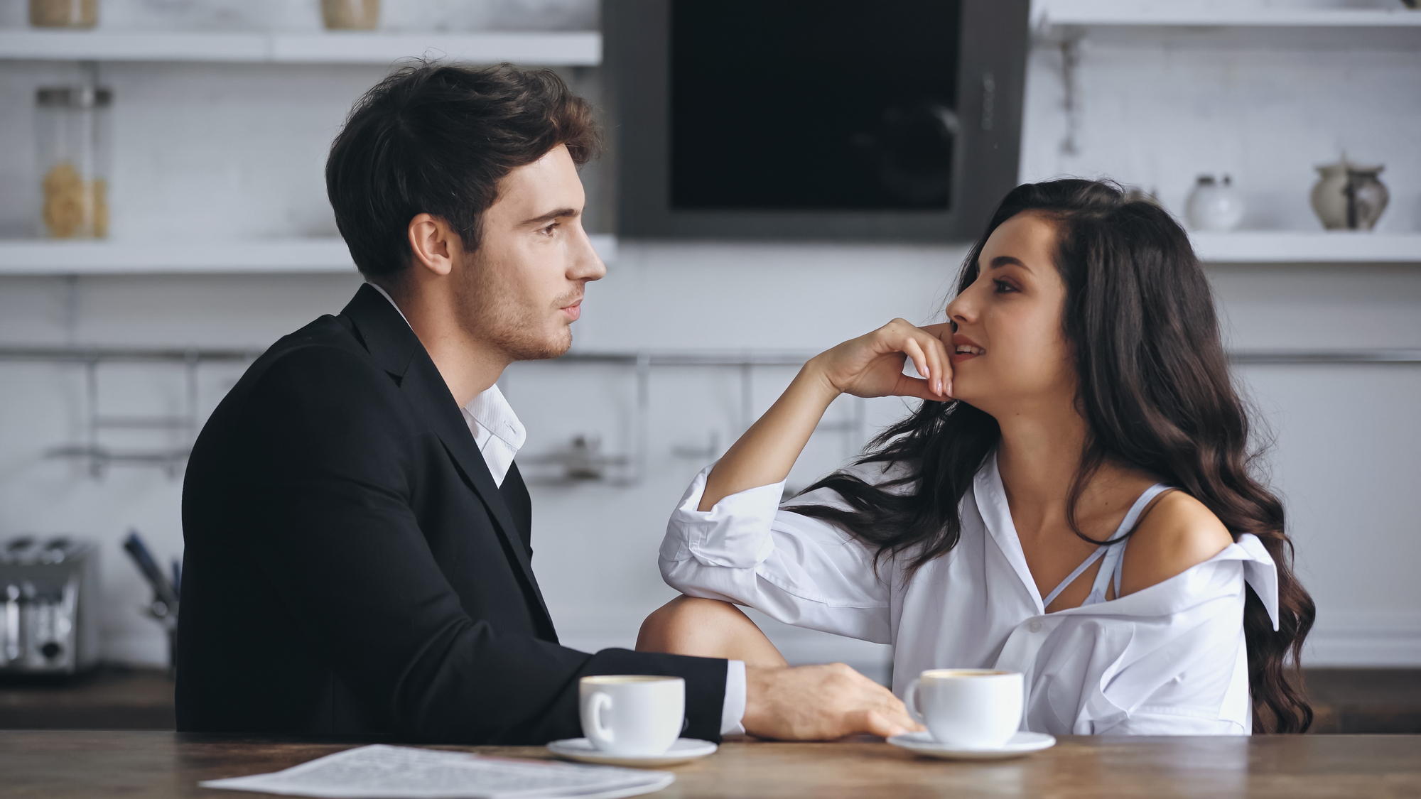 A man and woman sit at a cafe table, each with a cup of coffee. They are engaged in conversation, and the woman is smiling warmly while resting her head on her hand. The background shows a modern, minimalistic interior with shelves and a TV screen.