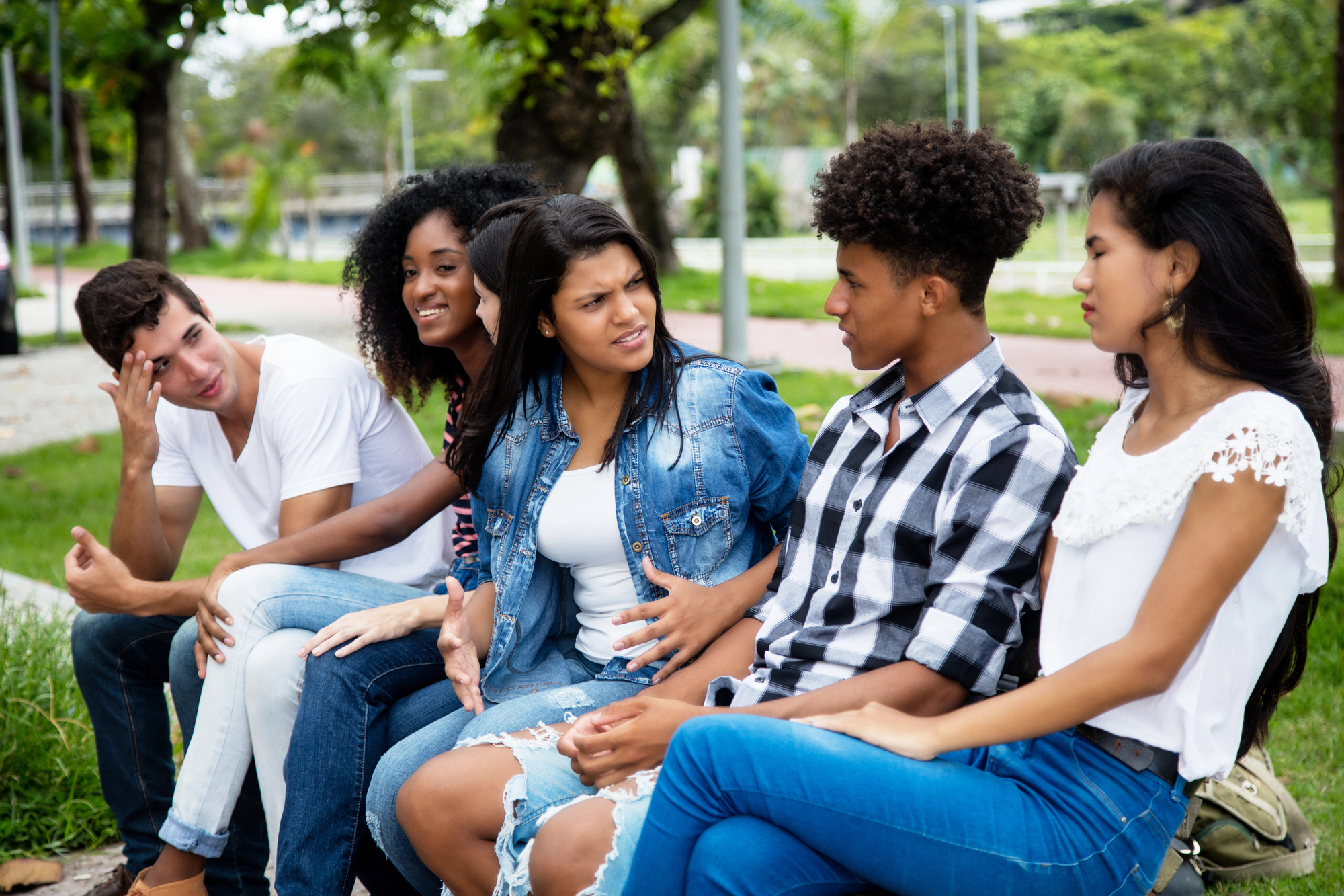 A group of five young adults is sitting on a park bench, engaged in conversation. They are casually dressed, and the background shows trees, grass, and a pathway. The individuals appear animated, with one gesturing while others listen and respond.