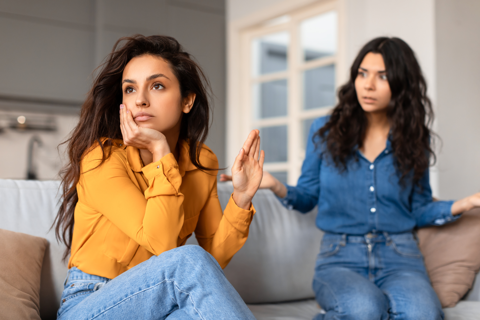 Two women sit on a couch in a domestic setting. The woman in the yellow shirt looks away with her hand raised, appearing uninterested or dismissive. The woman in the blue shirt, sitting behind her, appears to be speaking or reacting with concern or frustration.