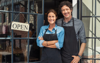 A smiling man and woman, both wearing black aprons, stand in the doorway of a store with an "OPEN" sign hanging on the glass door. The woman has her arms crossed, and the man stands with one hand in his pocket. They appear to be welcoming customers.