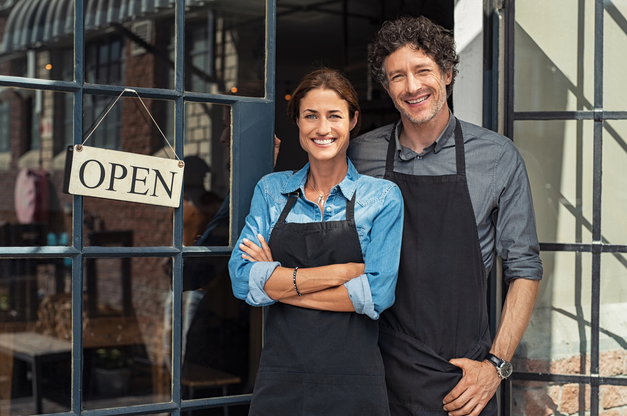 A smiling man and woman, both wearing black aprons, stand in the doorway of a store with an "OPEN" sign hanging on the glass door. The woman has her arms crossed, and the man stands with one hand in his pocket. They appear to be welcoming customers.