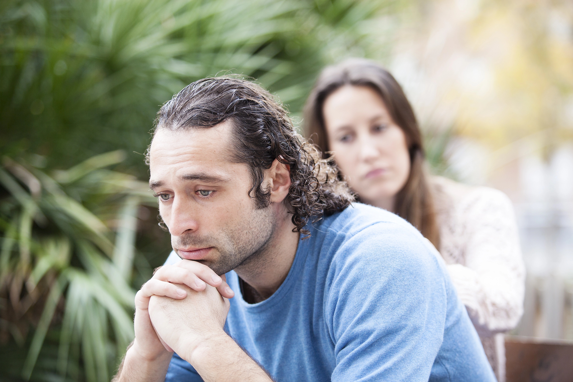 A man with long curly hair wearing a blue shirt sits pensively with his head resting on his hands, while a woman with long hair in the background gestures behind him. They appear to be outdoors, with green foliage visible in the background.