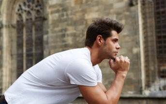 A man in a white T-shirt is leaning on a stone ledge and looking thoughtfully into the distance. The background features a historic, stone building with arched windows.