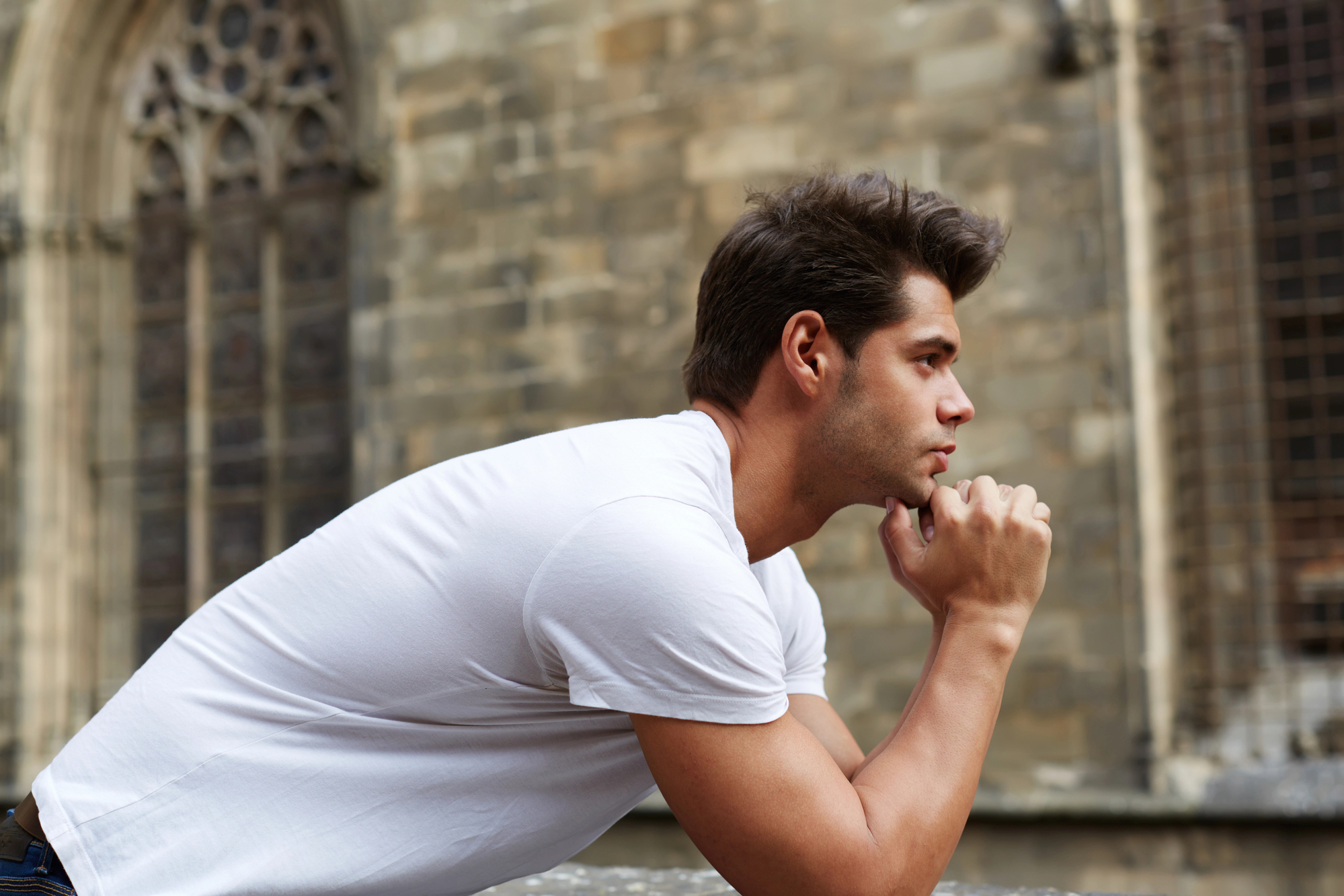 A man in a white T-shirt is leaning on a stone ledge and looking thoughtfully into the distance. The background features a historic, stone building with arched windows.