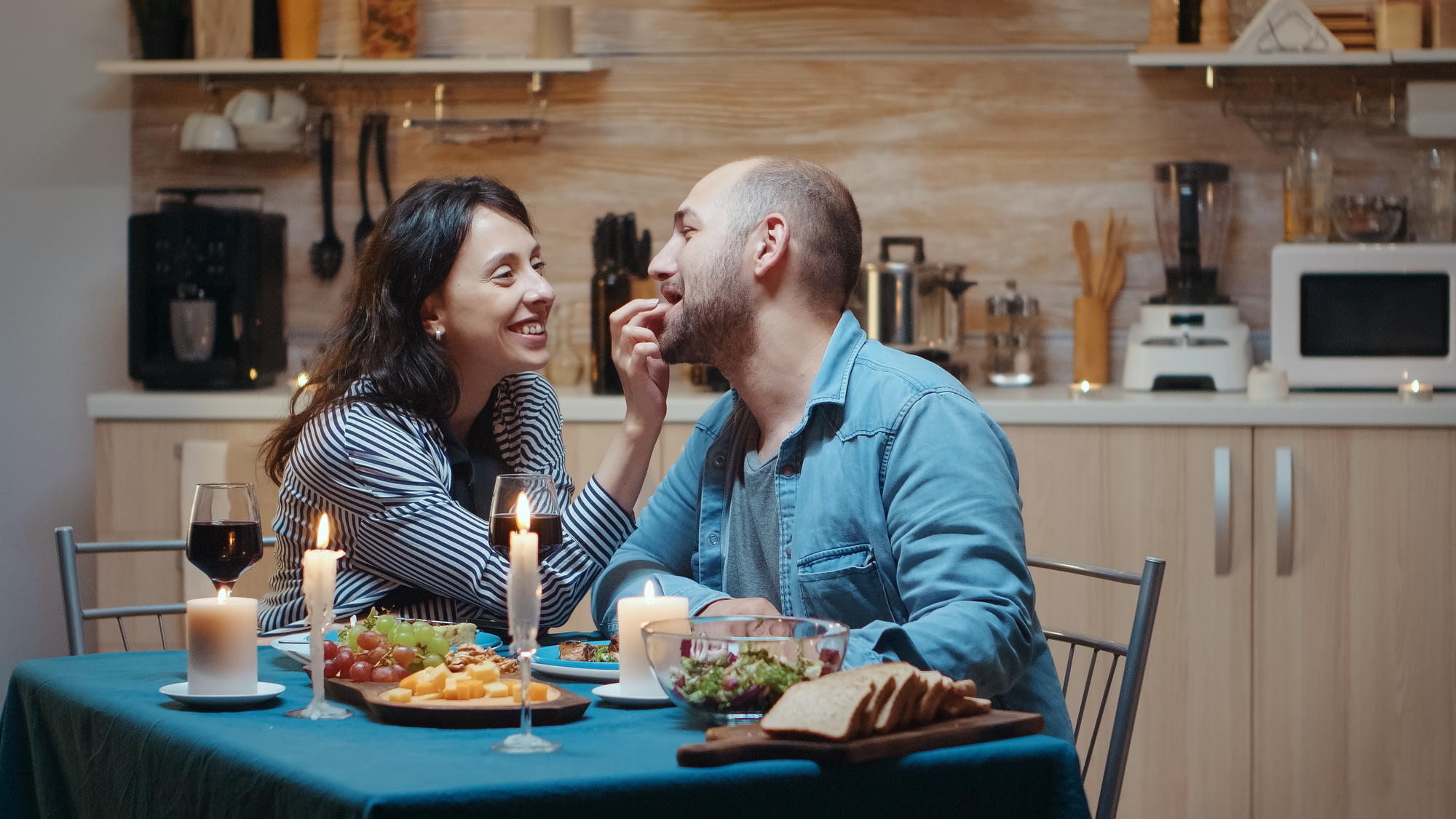 A happy couple sits at a dining table decorated with lit candles and a variety of foods, including bread, salad, and fruits. The woman, smiling, reaches across the table to touch the man's cheek, creating a warm and intimate ambiance in a cozy kitchen setting.