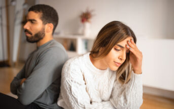 A man and a woman sit back-to-back on the floor, both appearing upset. The man, with a beard and wearing a gray sweater, looks away with arms crossed. The woman, in a white knitted sweater, leans forward with one hand on her forehead, looking distressed.