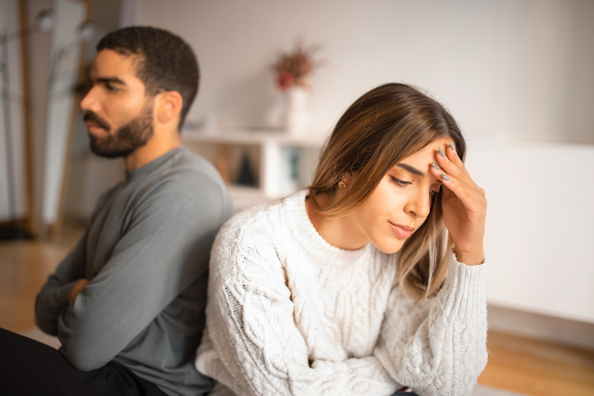 A man and a woman sit back-to-back on the floor, both appearing upset. The man, with a beard and wearing a gray sweater, looks away with arms crossed. The woman, in a white knitted sweater, leans forward with one hand on her forehead, looking distressed.