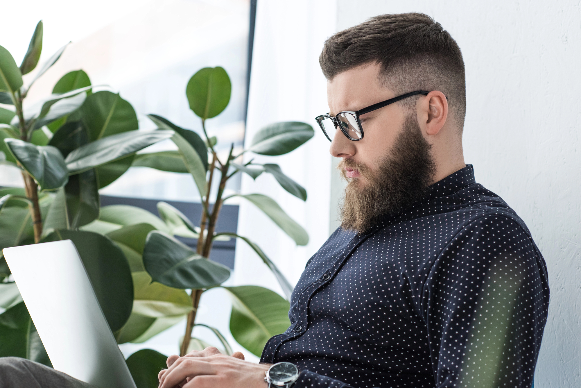 A bearded man wearing glasses and a dark polka-dot shirt is sitting and working on a laptop. He is seated by a window with broad-leafed plants in the background. The scene appears calm and focused.