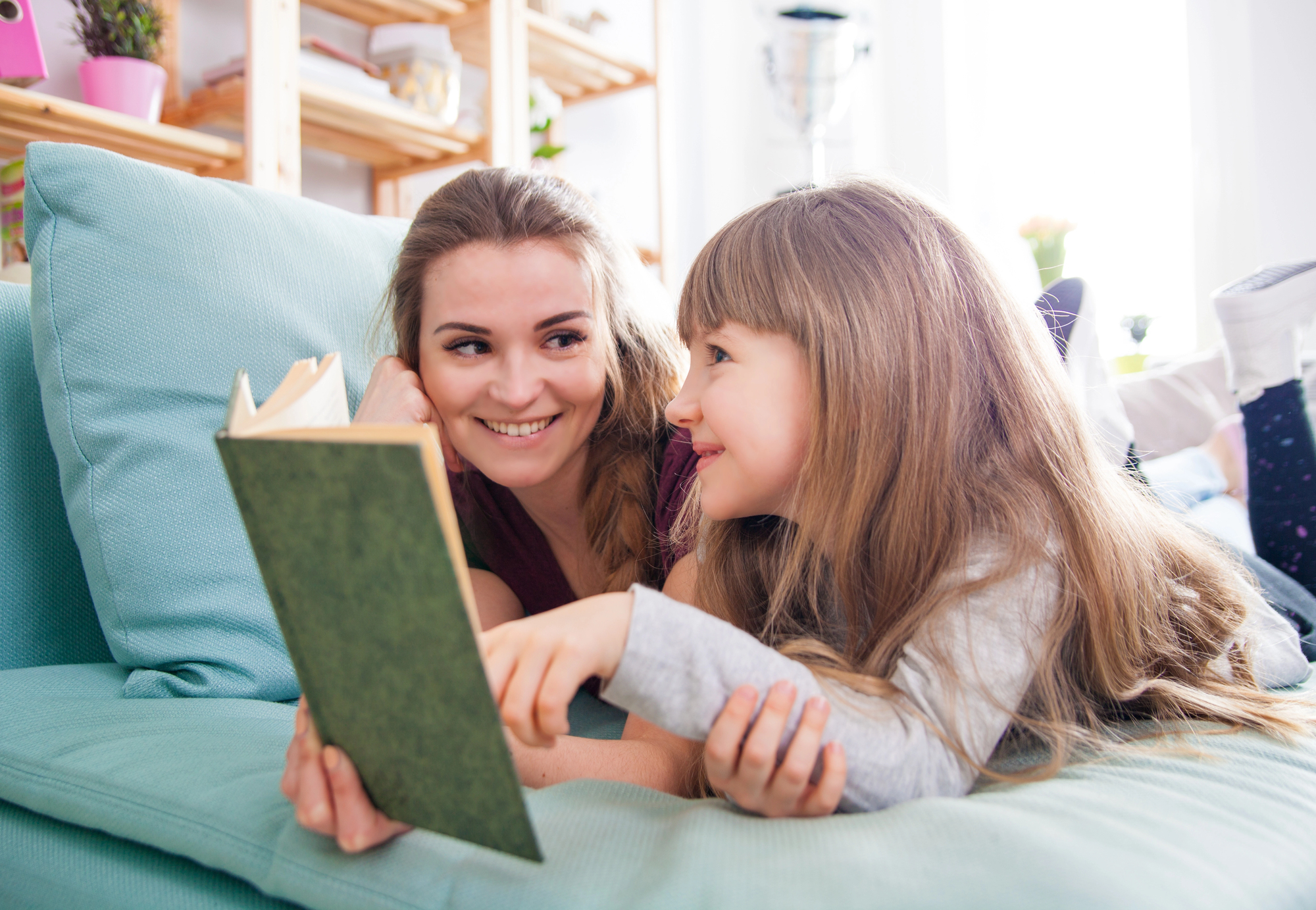 A woman and a young girl are lying on a couch, smiling at each other. The woman is holding an open book, and the girl is pointing at it. They appear to be enjoying a reading moment together. The background shows a cozy living space with shelves and various items.