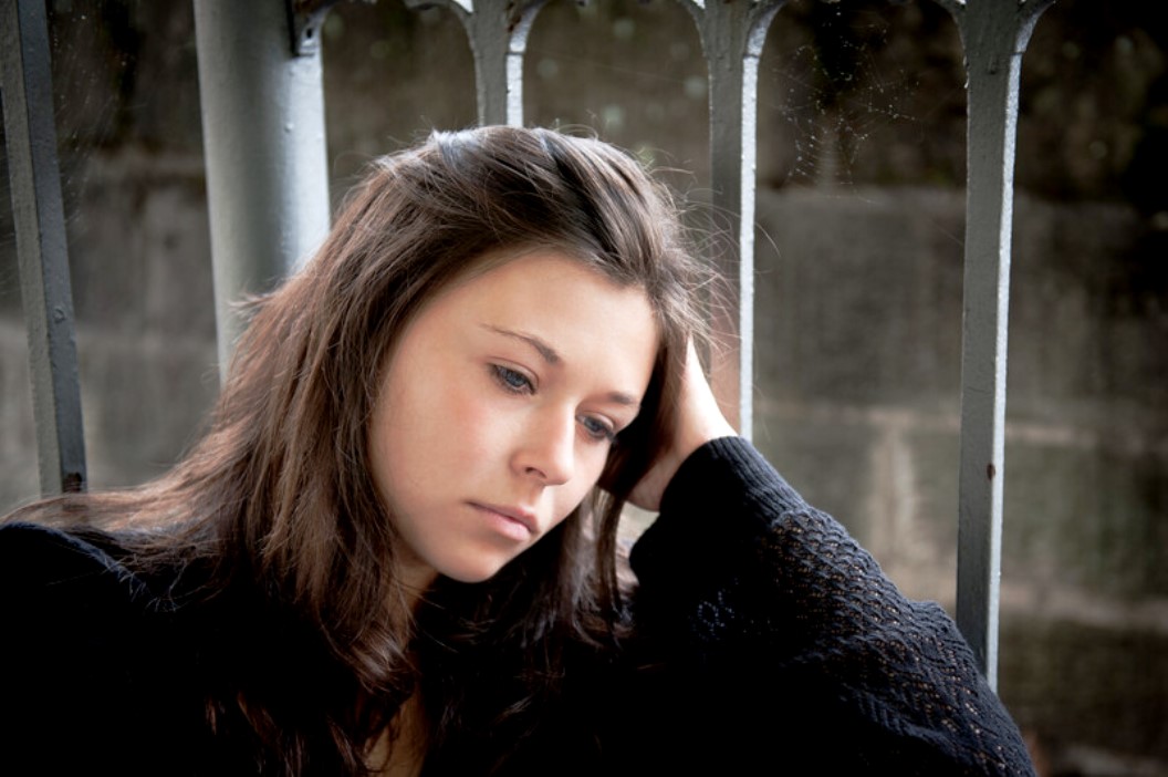 A young woman with long, dark brown hair is sitting while resting her head on her hand, looking off to the side with a pensive expression. She is wearing a dark long-sleeved top, and there is a gray metallic structure and a stone wall in the background.