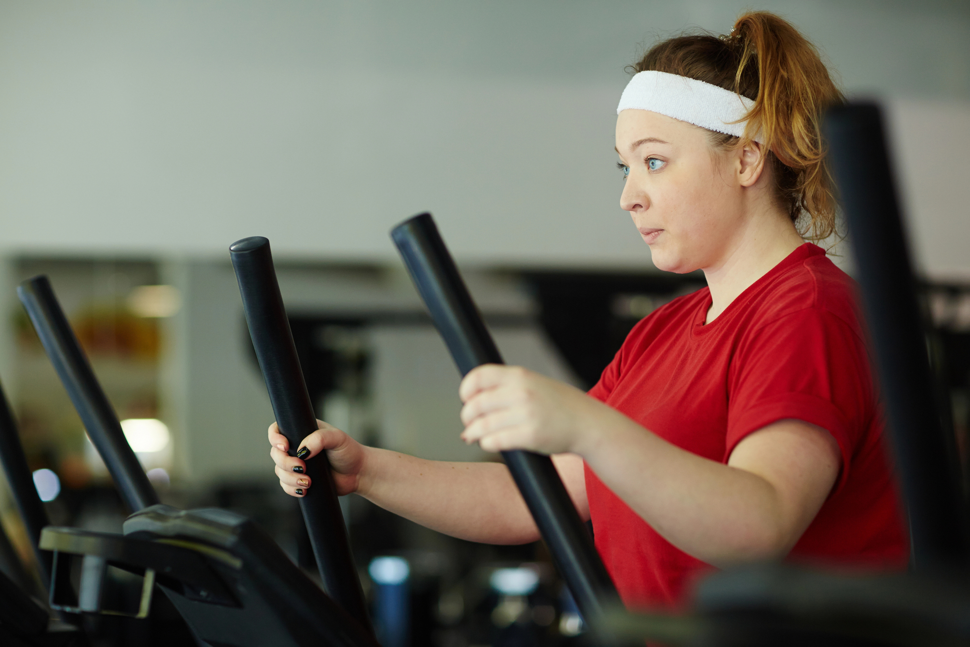 A woman wearing a red t-shirt and a white headband is using an elliptical machine at the gym. She has her hair tied back in a ponytail and is focused on her workout, with a look of determination on her face. The background shows gym equipment and a mirror.