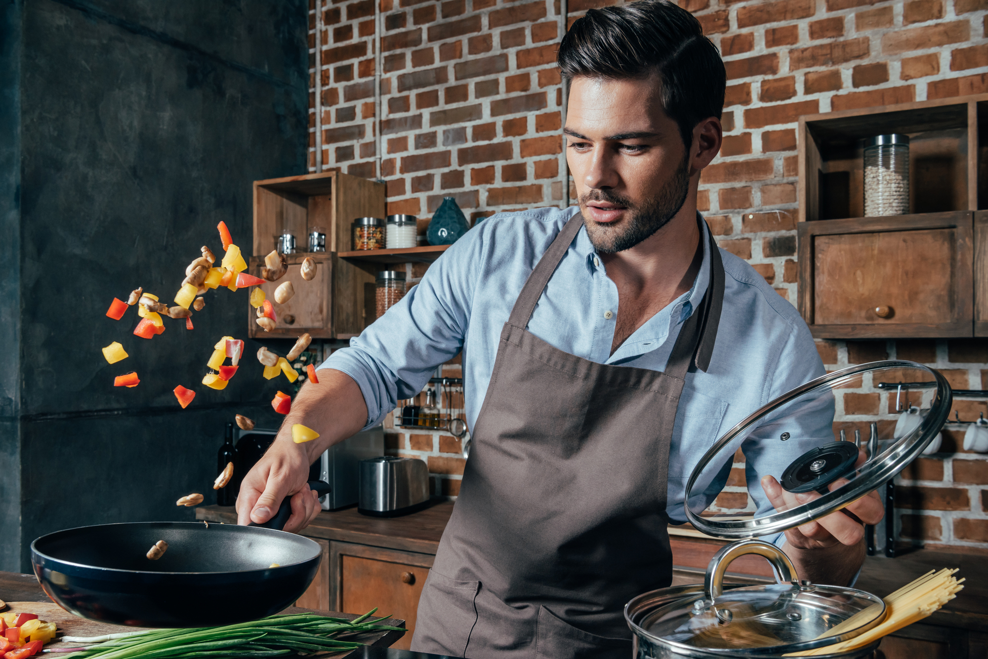 A man in a blue shirt and brown apron skillfully flips a frying pan with chopped vegetables, causing them to soar in the air. He holds a pot lid in the other hand. The kitchen has a rustic brick wall, wooden shelves, and various cooking utensils.