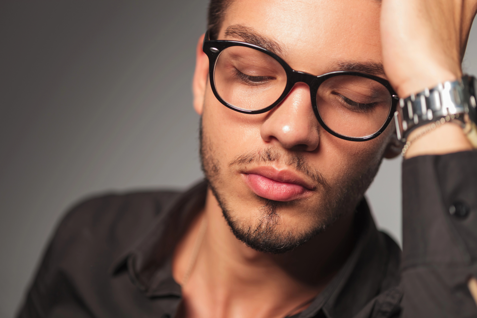 A man with short hair, glasses, and light facial hair is wearing a black shirt and a silver wristwatch. He is resting his head on his hand and appears to be in a contemplative or tired state, with his eyes closed and lips slightly parted.