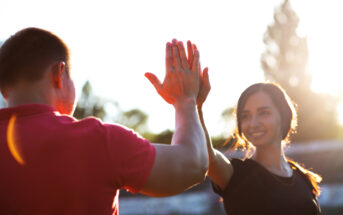 Two people high-fiving outdoors with the sun setting behind them. The person on the left is wearing a red short-sleeved shirt, and the person on the right is wearing a black short-sleeved shirt and smiling. Trees and bright sunlight are visible in the background.