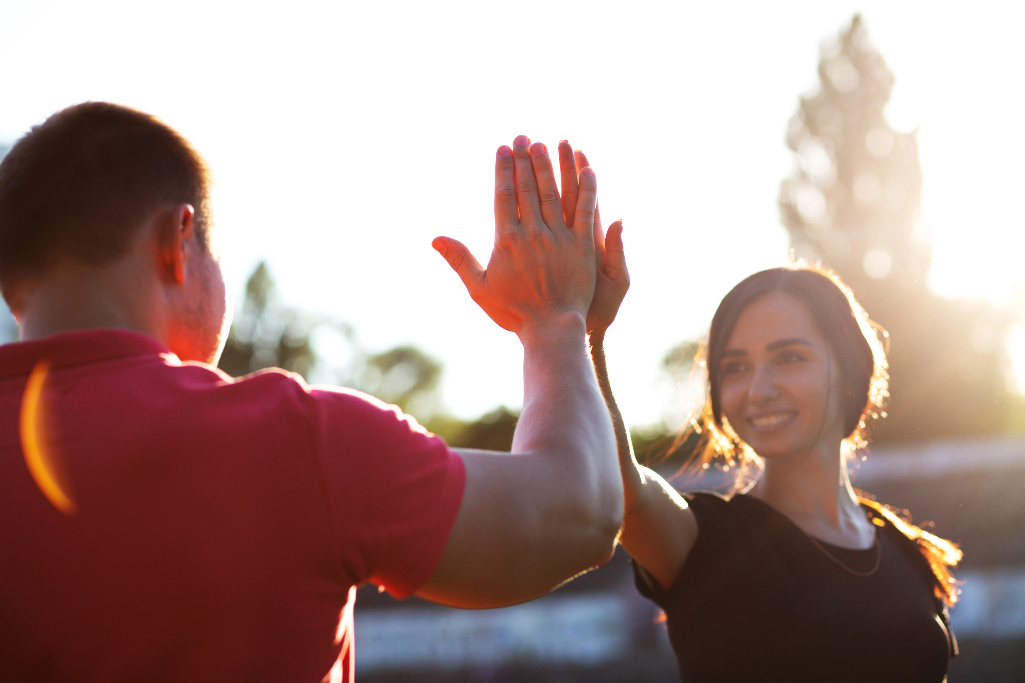 Two people high-fiving outdoors with the sun setting behind them. The person on the left is wearing a red short-sleeved shirt, and the person on the right is wearing a black short-sleeved shirt and smiling. Trees and bright sunlight are visible in the background.