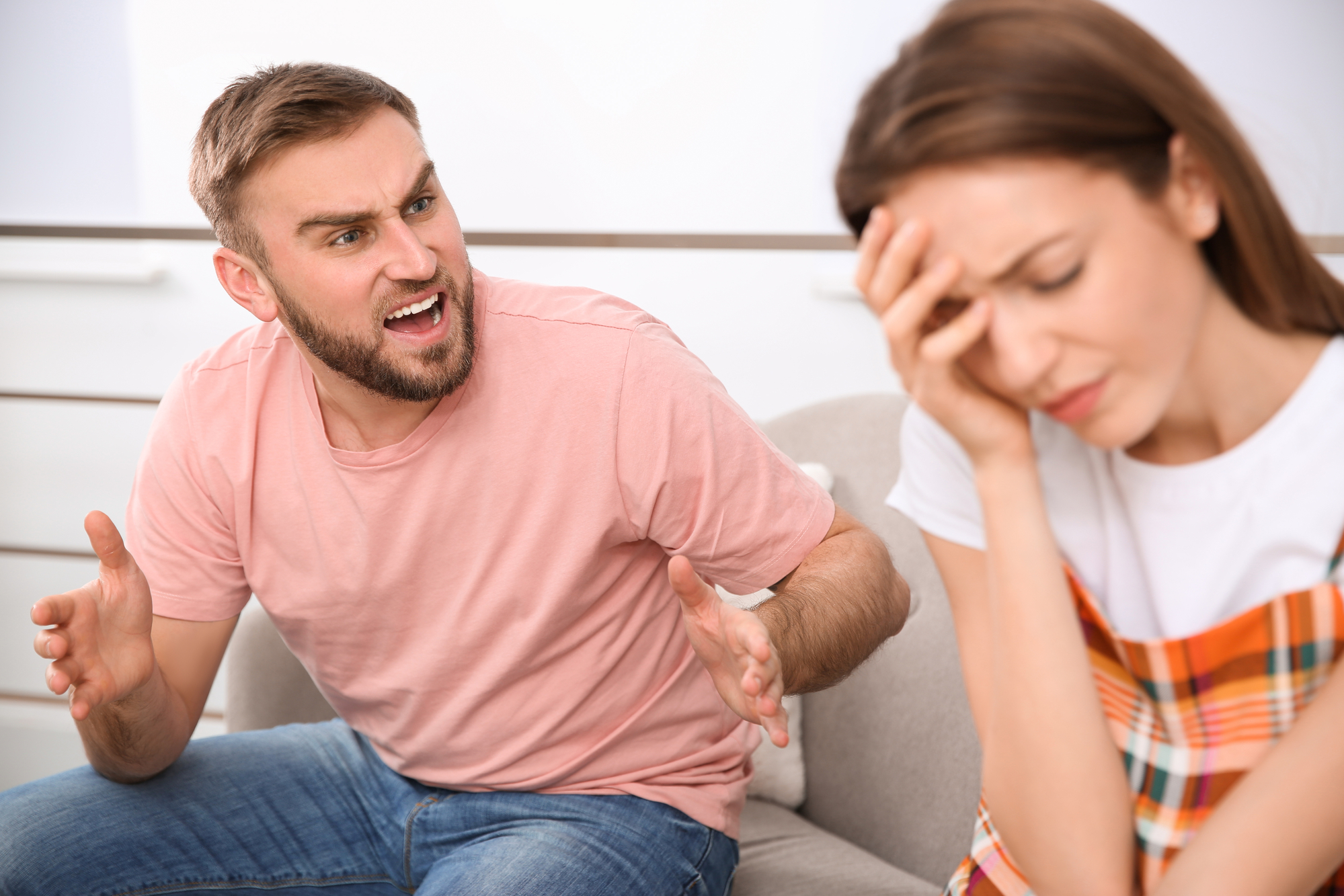 A man wearing a pink t-shirt is angrily arguing with a woman who appears distressed. The woman, wearing a plaid-patterned outfit, has her eyes closed and is holding her forehead with one hand, sitting on a sofa in a tense environment.