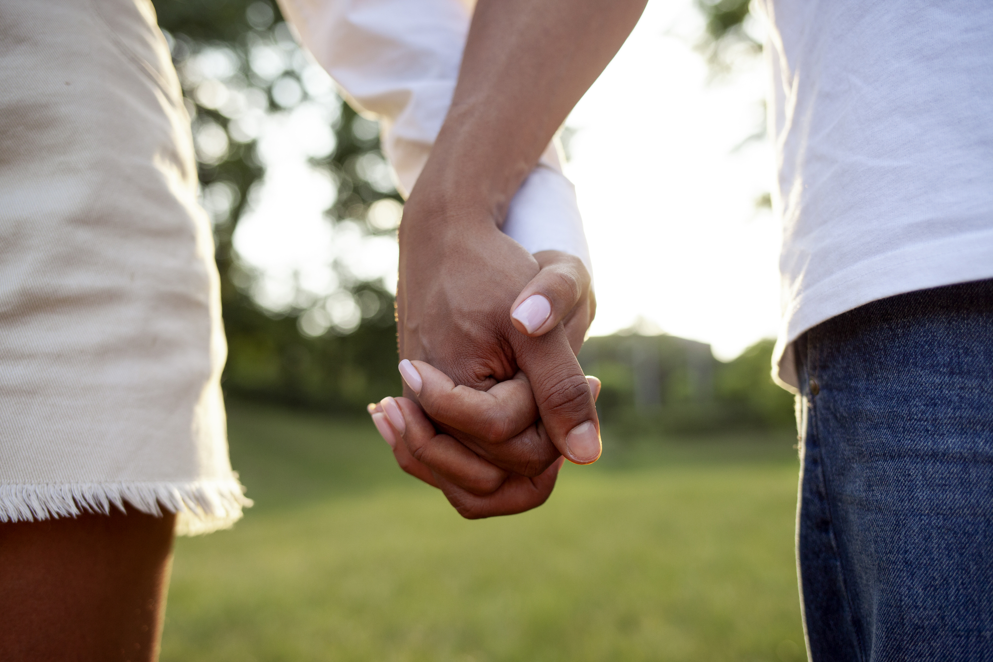 Two people are holding hands outdoors. One person wears a white skirt, and the other wears blue jeans. The background features greenery and is bathed in soft sunlight. The focus is on their clasped hands, symbolizing connection and affection.