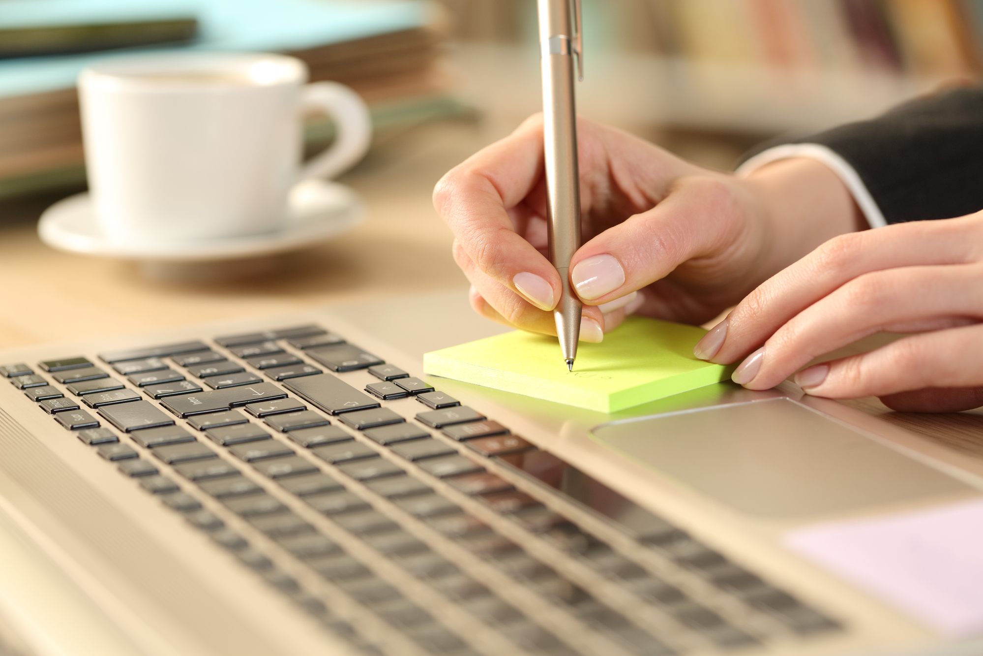 A person is working at a desk, holding a silver pen and writing on a yellow sticky note. Their other hand is resting on the keyboard of a laptop. A white cup sits in the background, out of focus.