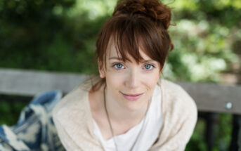 A woman with red hair and blue eyes is sitting outdoors on a wooden bench. She has her hair tied up in a messy bun and is wearing a light-colored top and a necklace. The background is blurred with greenery. She looks into the camera with a slight smile.